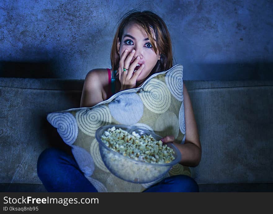 Young beautiful and scared hispanic woman watching horror suspense movie and eating popcorn sitting at home sofa couch late night in fear facial expression covering with pillow in grunge background. Young beautiful and scared hispanic woman watching horror suspense movie and eating popcorn sitting at home sofa couch late night in fear facial expression covering with pillow in grunge background