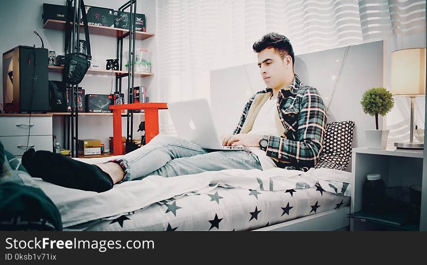 Man in Green, Blue, and Black Plaid Sports Shirt Sitting on Bed Using Silver Macbook