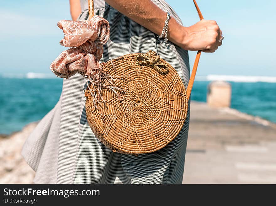 Woman Wearing Grey Skirt and Round Brown Rattan Crossbody Bag on Wooden Dock Near Body of Water