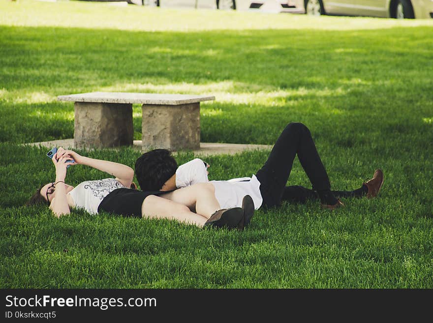 Man And Woman Laying On Green Grass Near Concrete Bench