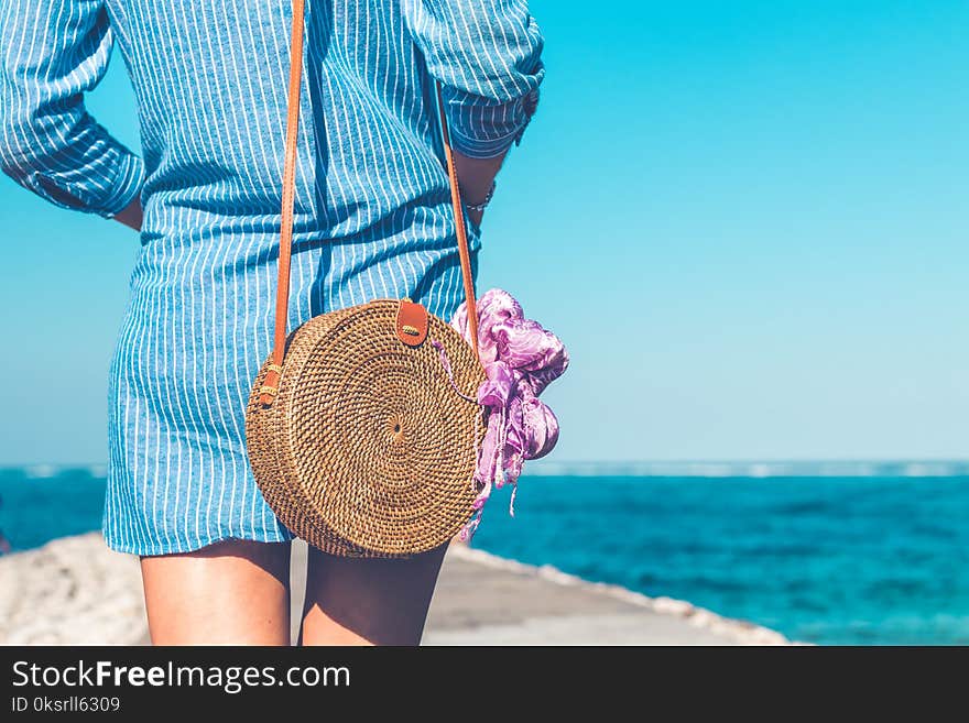 Woman Wearing Blue and White Striped Dress With Brown Rattan Crossbody Bag Near Ocean