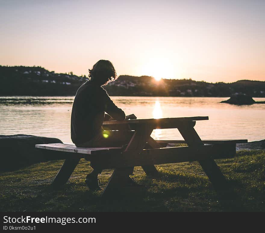 Silhouette of Person in Black Top Sitting on Picnic Bench Near Body of Water during Sunset