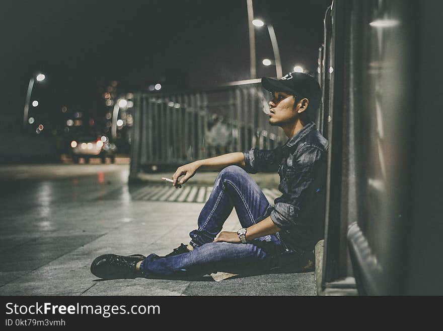 Shallow Focus Photography of Man Sitting Beside Railing Holding Cigarette Stick