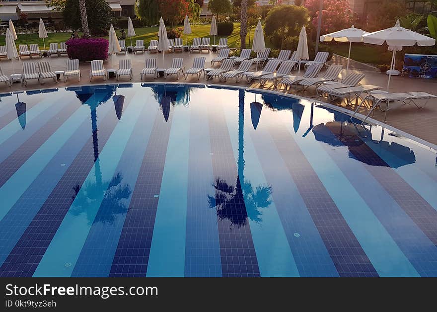 Reflection in the clear water of the swimming pool in luxury resort hotel. Summer vacation landscape. Sunset light and beautiful resort pool with nobody. Reflection of the palm trees in the pool water. Blue striped pool bottom and clear pool water surface at the sunshine. Tourism and vacation themed. Travel background. Deck chairs and white beach umbrellas near hotel pool. Reflection in the clear water of the swimming pool in luxury resort hotel. Summer vacation landscape. Sunset light and beautiful resort pool with nobody. Reflection of the palm trees in the pool water. Blue striped pool bottom and clear pool water surface at the sunshine. Tourism and vacation themed. Travel background. Deck chairs and white beach umbrellas near hotel pool.