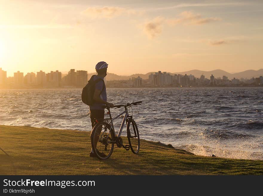 Man With Bicycle Standing on Cliff Front of Water Waves Golden Hour Photography