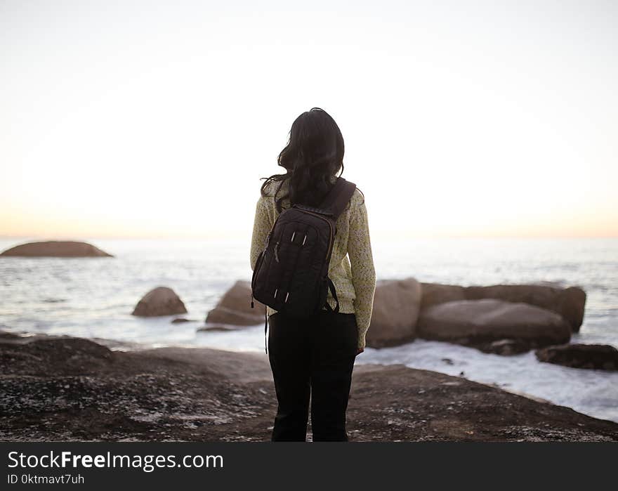 Woman Wearing White Long-sleeved Top and Black Pants Carrying Black Backpack While Standing on Shore during Adytime