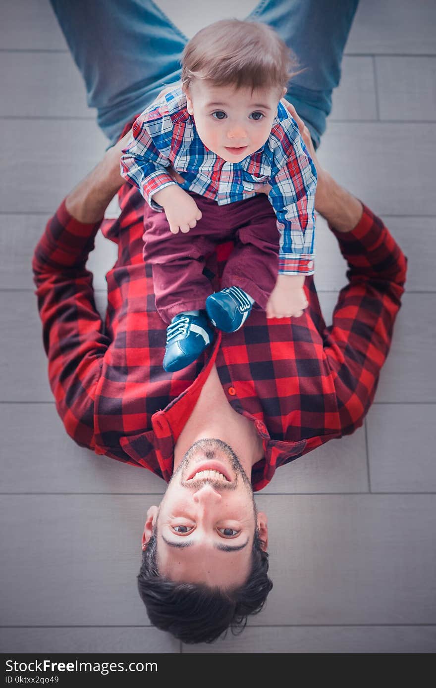 Lying Man Wearing Red and Black Gingham Long-sleeved Shirt Holding Toddler Sitting on His Stomach