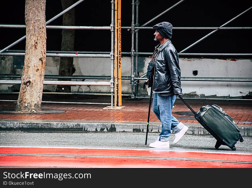 Walking Man in Black Leather Jacket and Blue Denim Pants Holding Luggage Bag