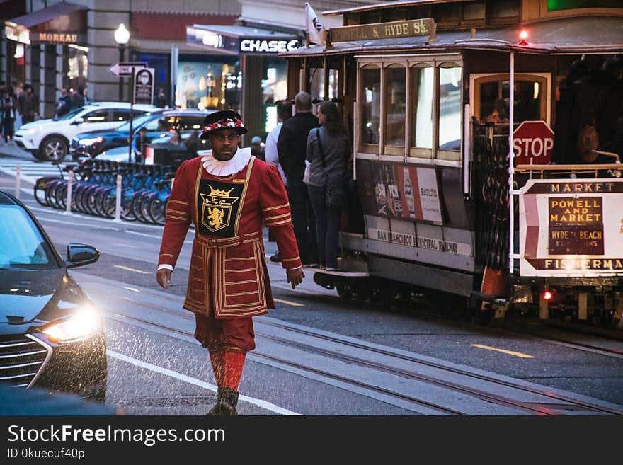 Man Wearing Red and Yellow Dress Walking on Concrete Road Near White and Red Cable Train