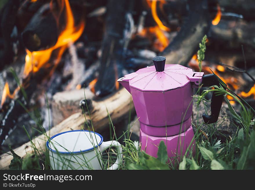 A mug and a geyser coffee machine near the bonfire. Camping in the forest. A cup stands on the grass against the background of a fire. A mug and a geyser coffee machine near the bonfire. Camping in the forest. A cup stands on the grass against the background of a fire.