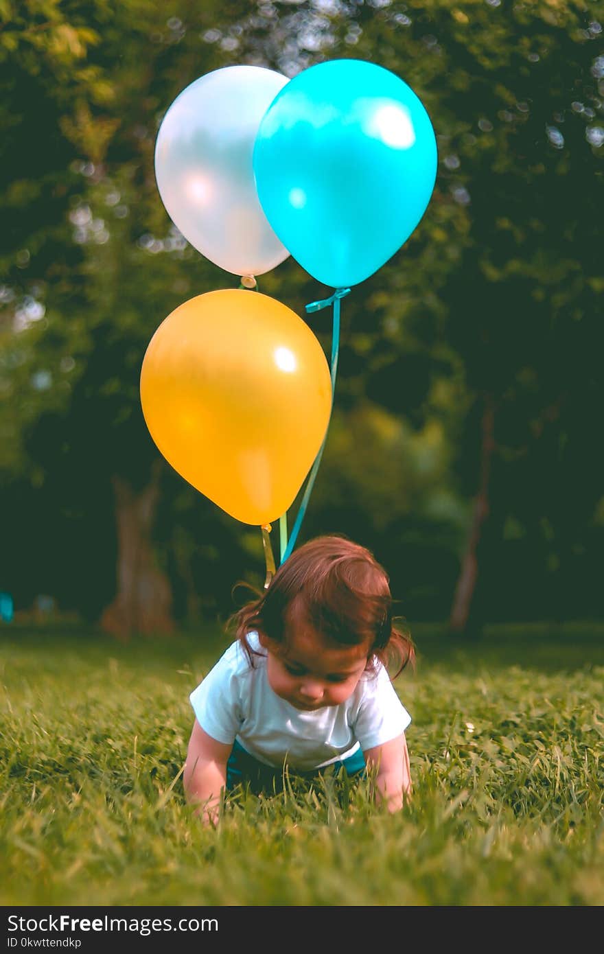 Baby Wearing White T-shirt Holding Three Yellow, Blue, and White Balloons on Green Grass Near Woods