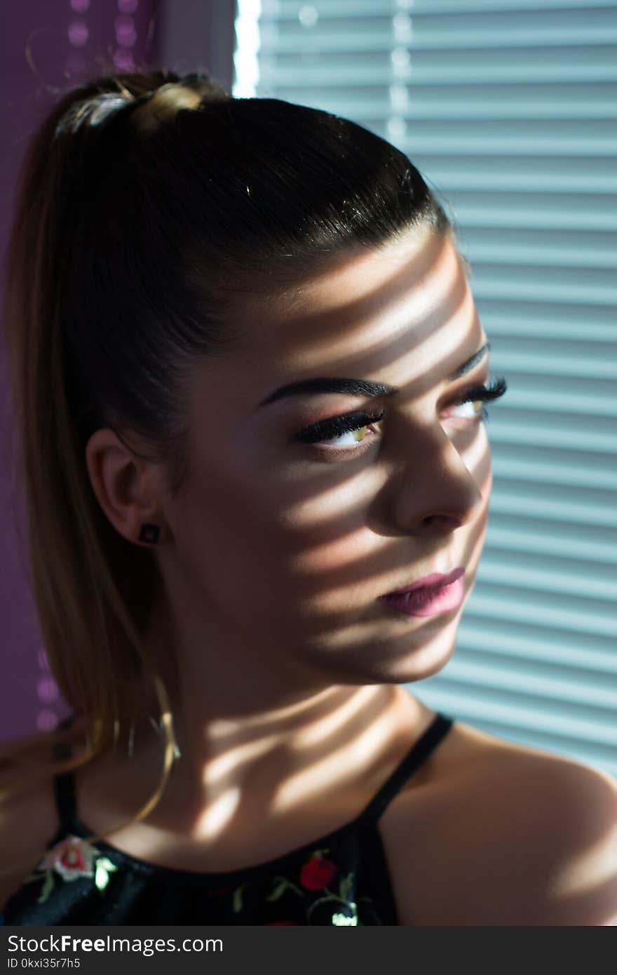 Serious girl face with shadow of Venetian blinds looking through window in sunset at home