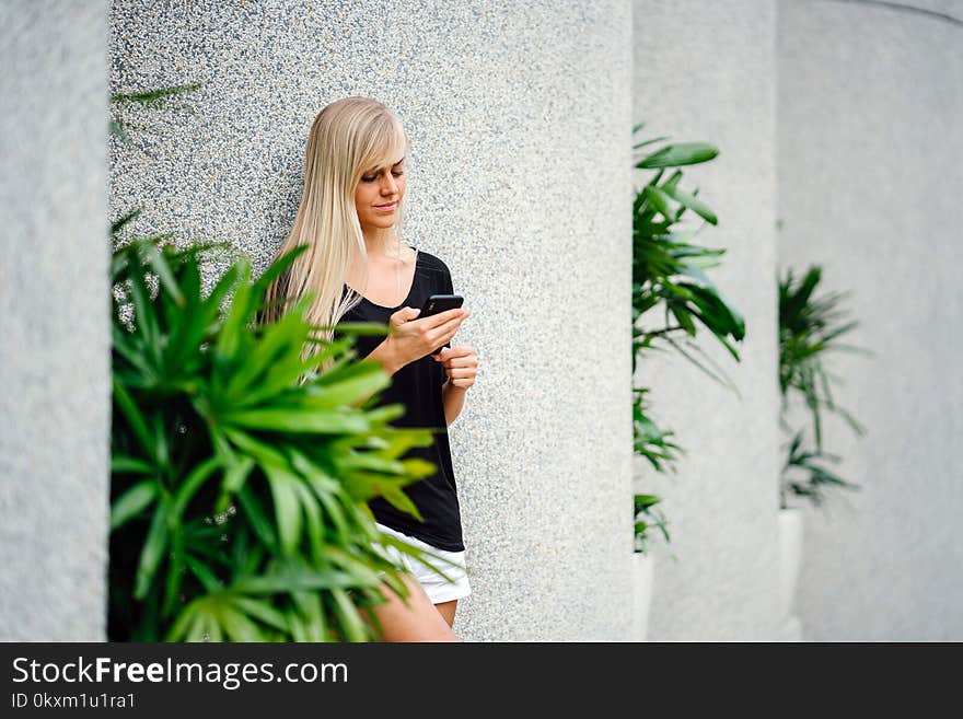 Woman Wearing Black Scoop-neck Shirt Standing in Front on Concrete Column Holding Smartphone