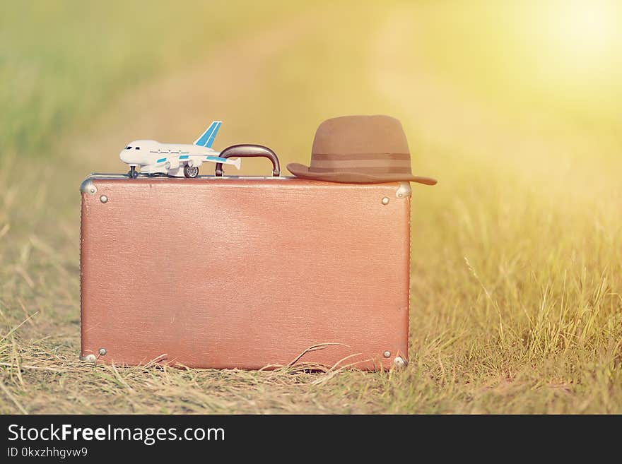 Travel and adventure concept. Vintage brown suitcase and hat with toy airplane near the road in the green field.