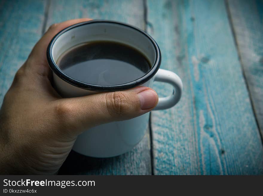 man holding in hand white iron mug with coffee or black tea on an old blue wooden table