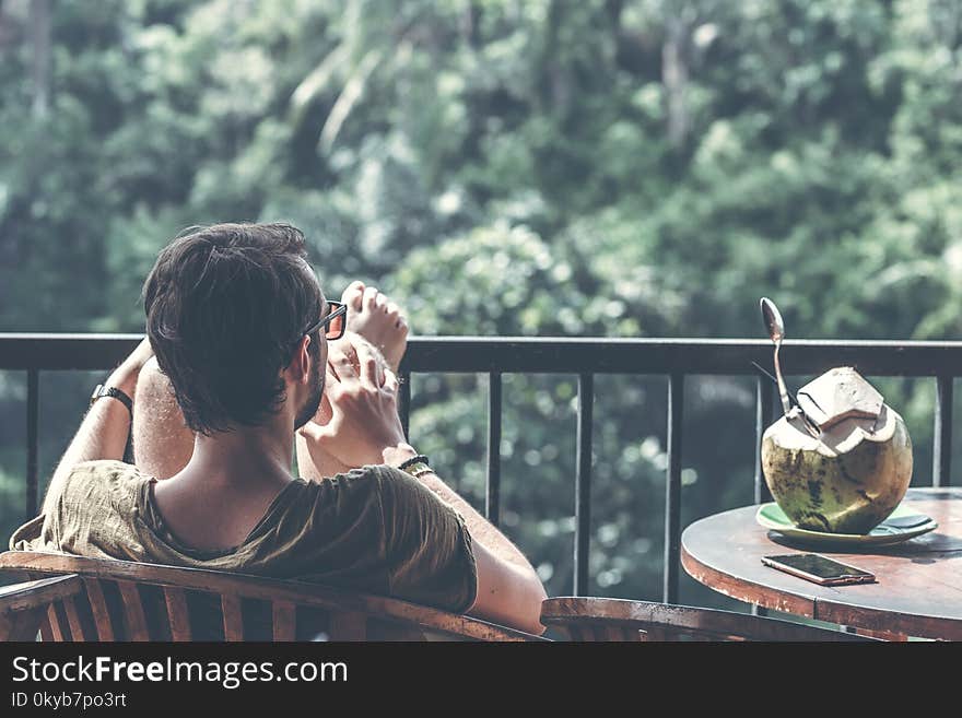 Man Sitting on Wooden Chair With Foot on Black Steel Railings