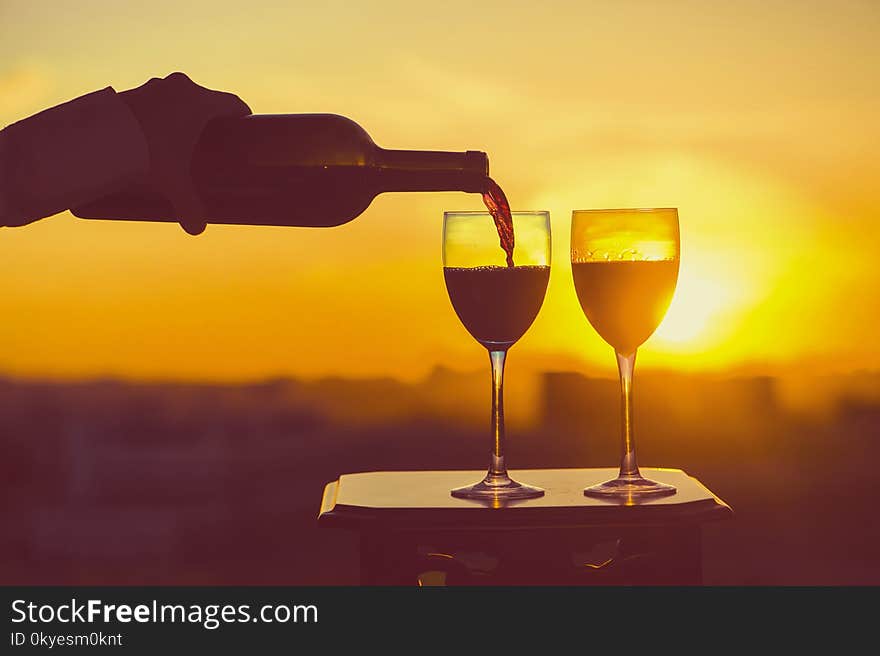 Female hand with bottle pours red wine into glasses on a sunset background.