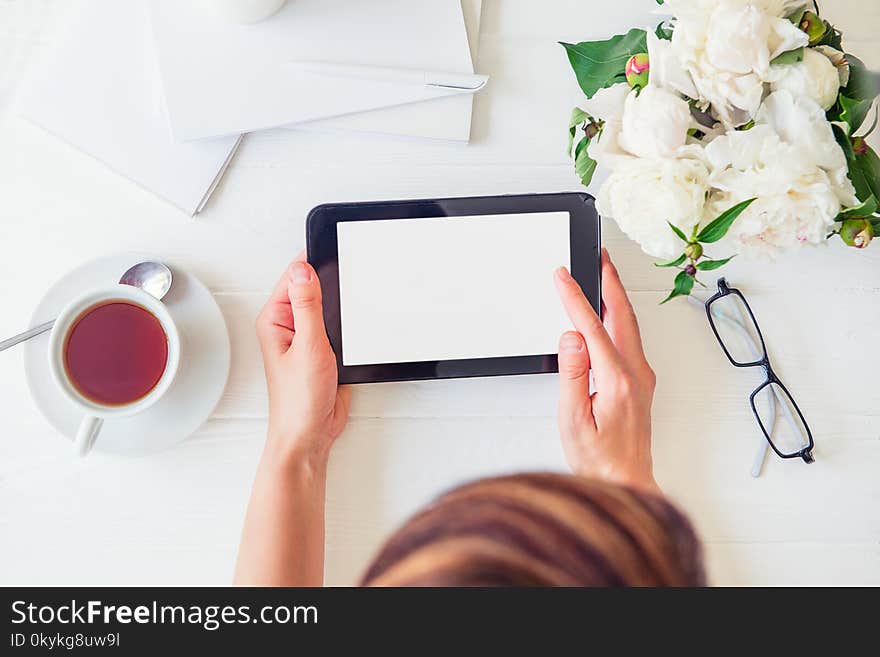 Workspace with girl`s hands holding tablet with blank screen, cup of tea, glasses, notebooks, white peony on woodden table. Top view feminine office desk. Planning day. Freelancer working place