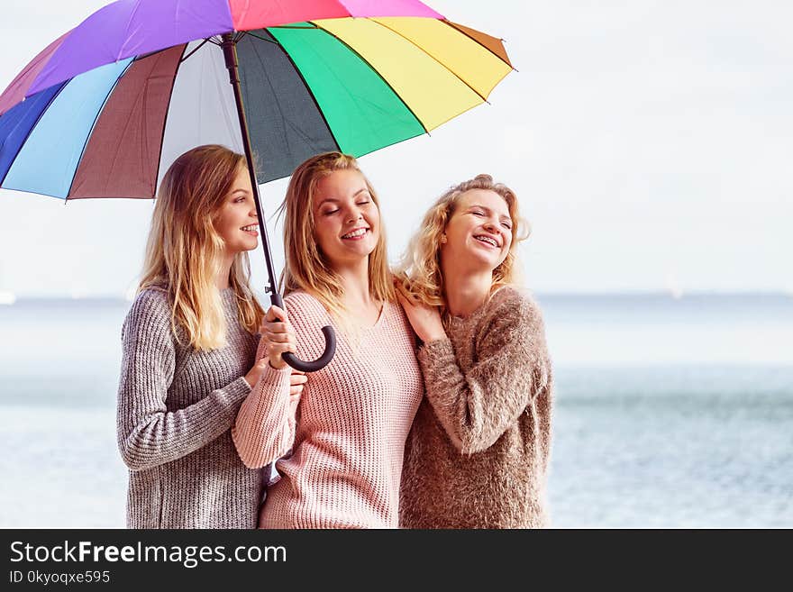 Three pretty young women friends under colorful umbrella parasol. Fashionable females wearing sweaters spending time outdoor. Three pretty young women friends under colorful umbrella parasol. Fashionable females wearing sweaters spending time outdoor.