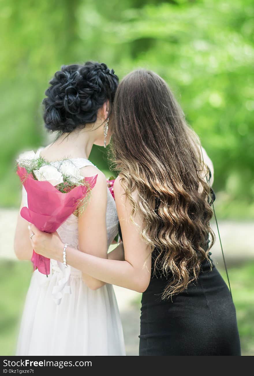 Portrait of back of two young beautiful women hugging each other in green summer park. Pretty females Bride and bridesmaid talking.