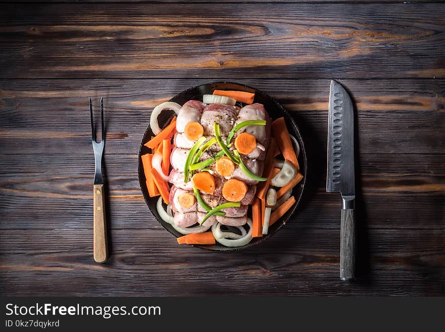 Raw pork chop marinated meat roll with coarse sea salt pepper assorted vegetables with carrots green onions and onion rings in cast iron skillet On wooden table background served with meat fork and knife. Raw pork chop marinated meat roll with coarse sea salt pepper assorted vegetables with carrots green onions and onion rings in cast iron skillet On wooden table background served with meat fork and knife.