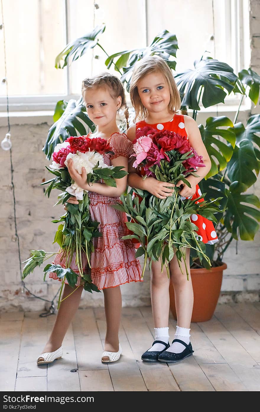 Two cute little girls with peonies. Two beautiful friends having fun, hugging and smiling inside at home or in the studio