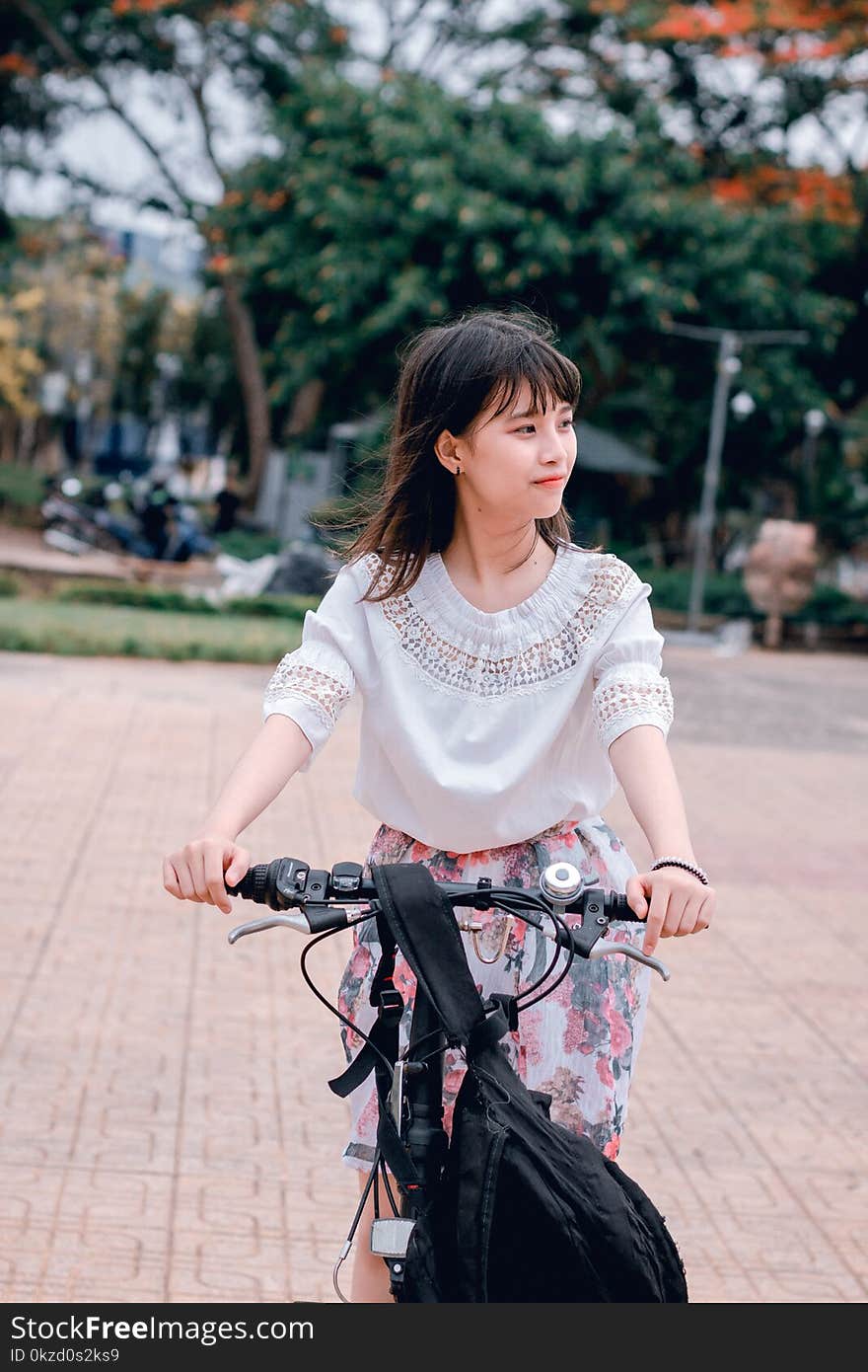 Woman Wearing White Blouse Riding Bicycle on Brown Concrete Tiled Area Near Trees