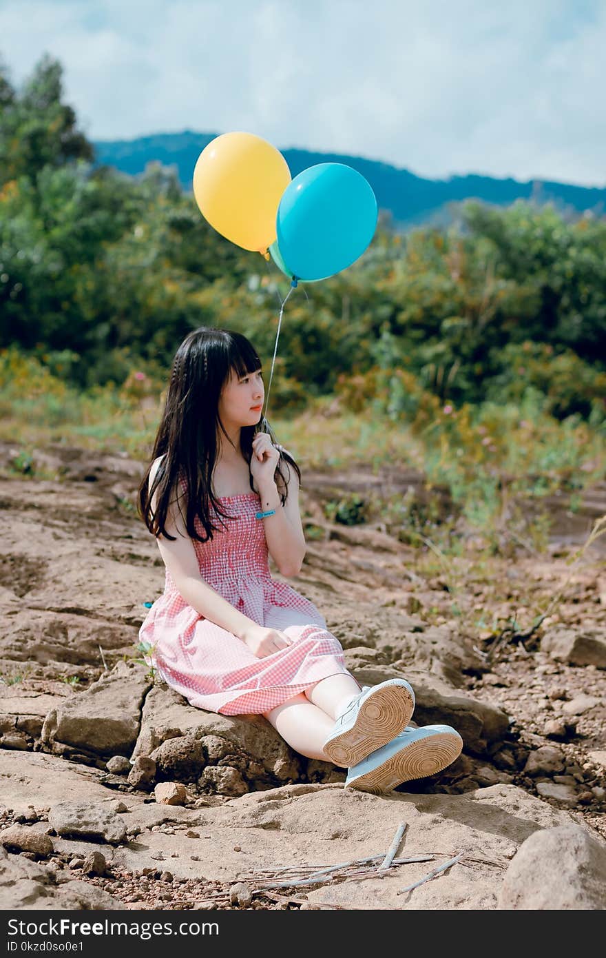 Woman Wearing Pink Dress Sitting on Ground Holding Two Balloons