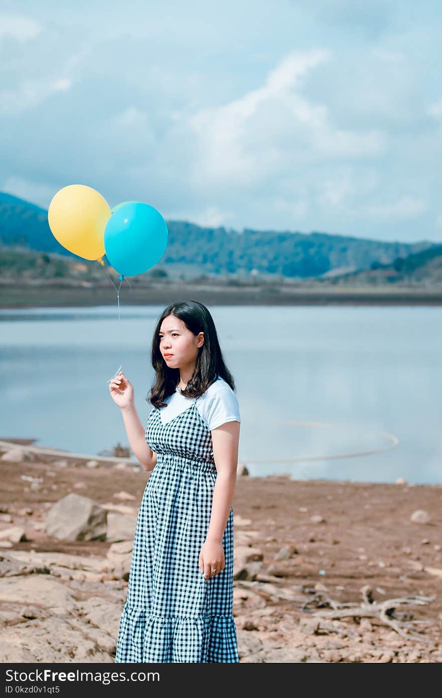 Woman Wearing Blue and White Plaid Dress Holding Blue and Yellow Balloons Near Calm Body of Water at Daytime