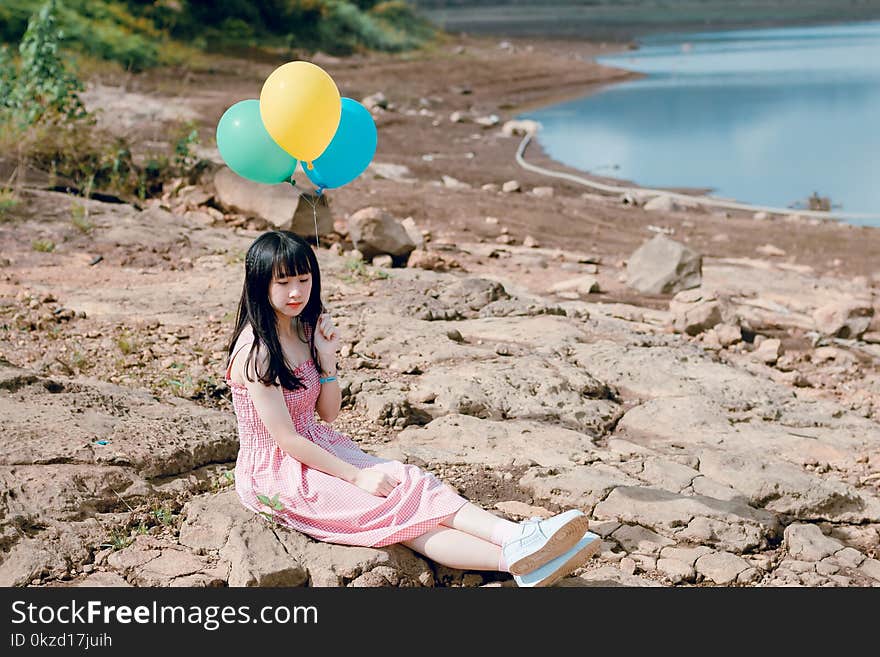 Woman in Pink Thick Strap Dress Holding a Yellow and Blue Balloons While Sitting on Rock Terrain