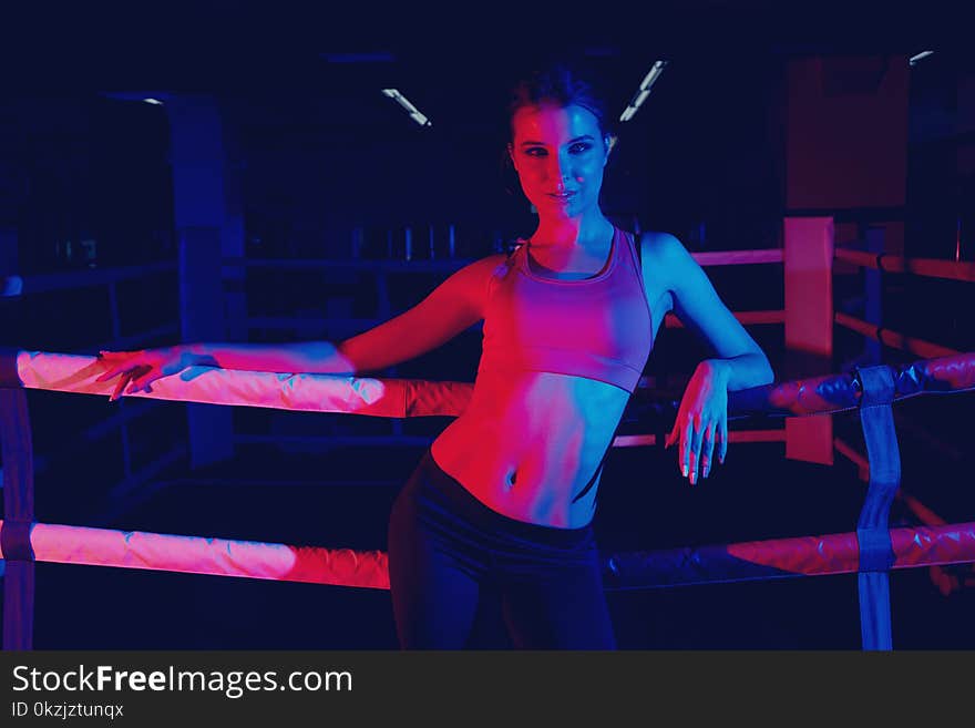 Attractive brunette woman with pony tail, dressed in sportswear, illuminated by dim neon light, sitting and leaning her back against ropes of boxing ring. Beautiful female fitness model posing at gym.