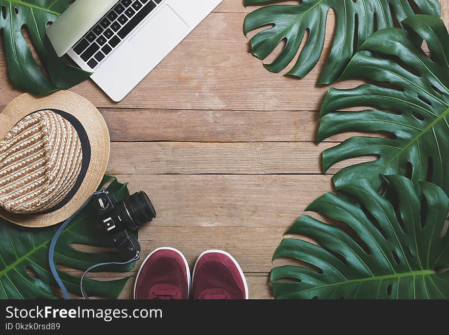 Flat lay creative frame of tropical nature leaves Monstera on rustic wood grunge background with laptop computer, retro camera, hat and pair of sneaker shoes, digital nomad and life balance concepts.