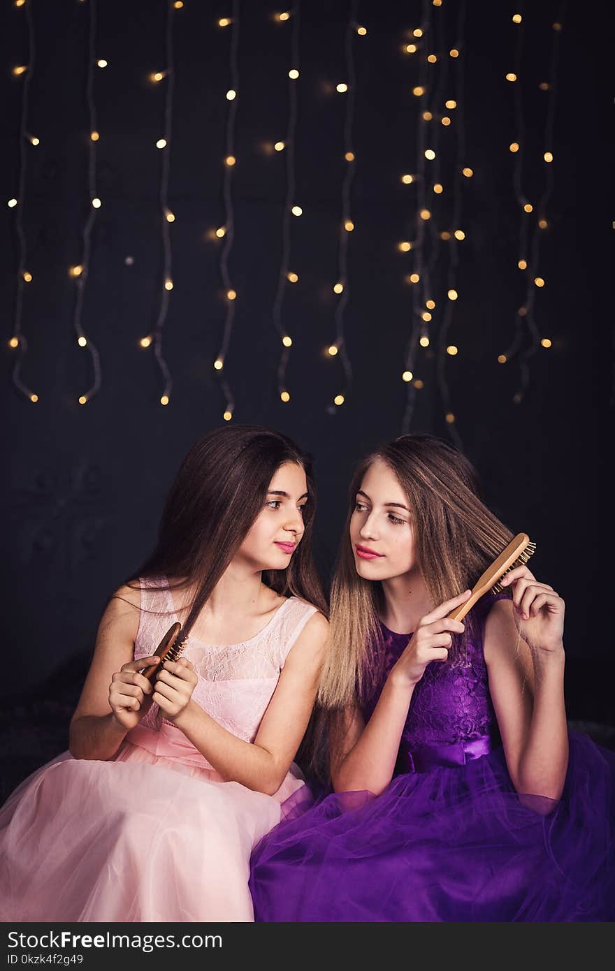 Cheerful girlfriends. Two girls comb each other`s hair, sitting side by side in studio on dark background. Copy space.