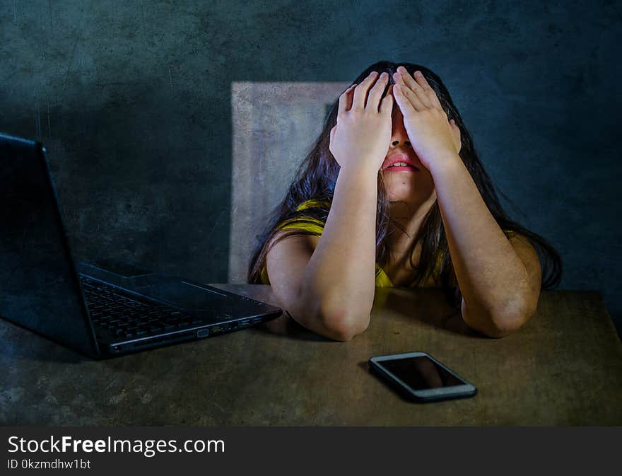 Dramatic portrait of young latin sad and scared woman covering face with hands desperate and depressed with laptop and mobile phone in cyber bullying and internet problem. Dramatic portrait of young latin sad and scared woman covering face with hands desperate and depressed with laptop and mobile phone in cyber bullying and internet problem