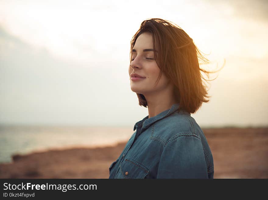 Great pleasure. Charming calm young woman is standing near sea with closed eyes and expressing delight. She is posing against wonderful sunset while enjoying last rays of the sun. Great pleasure. Charming calm young woman is standing near sea with closed eyes and expressing delight. She is posing against wonderful sunset while enjoying last rays of the sun