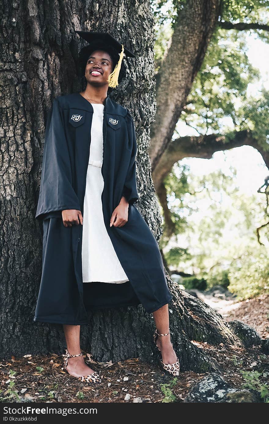 Smiling Woman Wearing Black Academic Gown and Hat Leaning Behind Tree