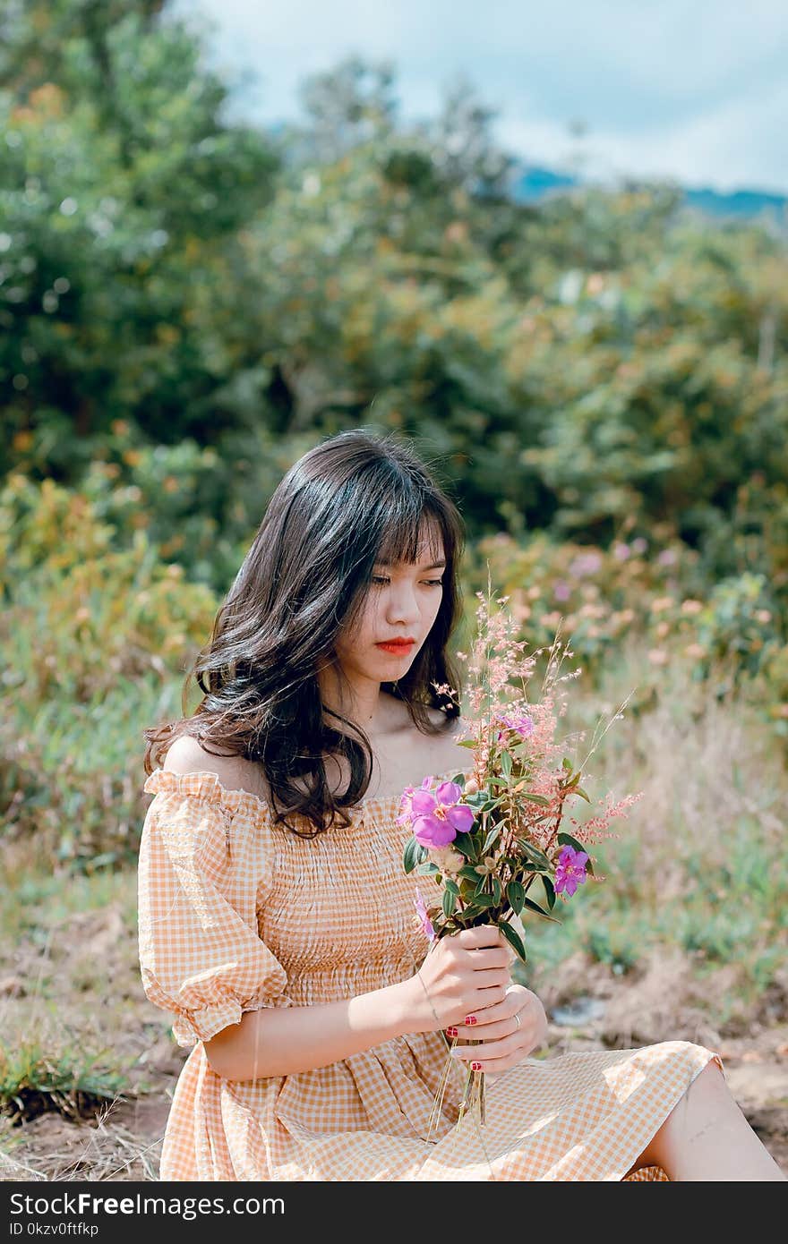 Shallow Focus Photography of Woman Wearing Orange Checked Dress Holding Purple Flowers