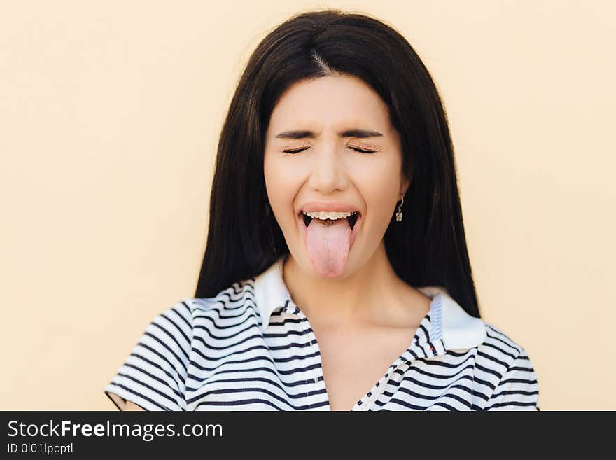 Portrait of beautiful woman makes grimace, keeps mouth wide opened and shows tongue, keeps eyes closed, wears casual clothes, poses against studio background. People and facial expressions concept.