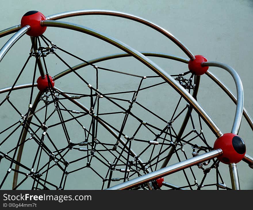 Red plastic ball joints with black vinyl rope mesh on a playground climbing equipment and shiny stainless steel tube framing and aluminum clamps in a public park under blue sky. Red plastic ball joints with black vinyl rope mesh on a playground climbing equipment and shiny stainless steel tube framing and aluminum clamps in a public park under blue sky