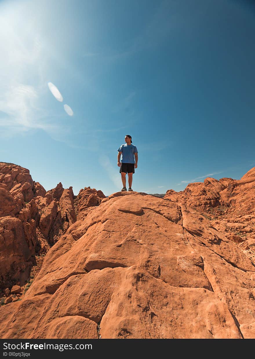 Man Wearing Blue Shirt and Black Shorts Standing on Top of Brown Rock Formations Under Clear Sky