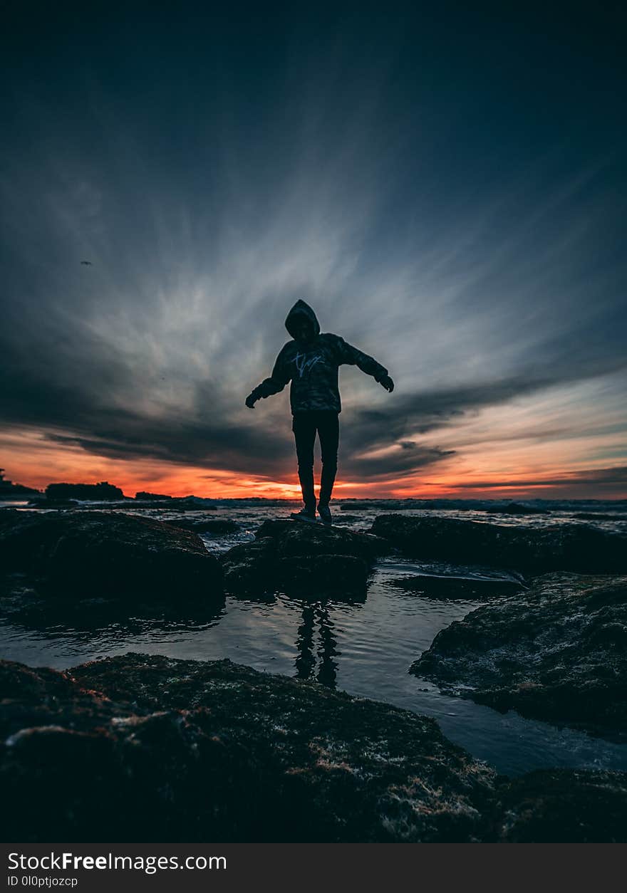 Person Wearing Hoodie Standing on Rock Surrounded by Body of Water