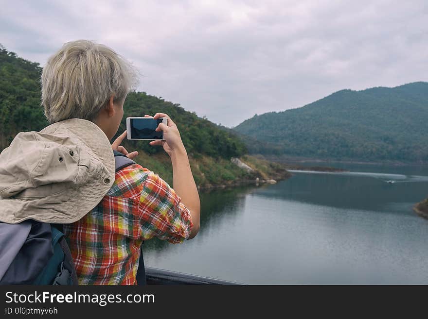 Person Holding Smartphone While Taking Photo of Calm Water Near Mountain at Daytime