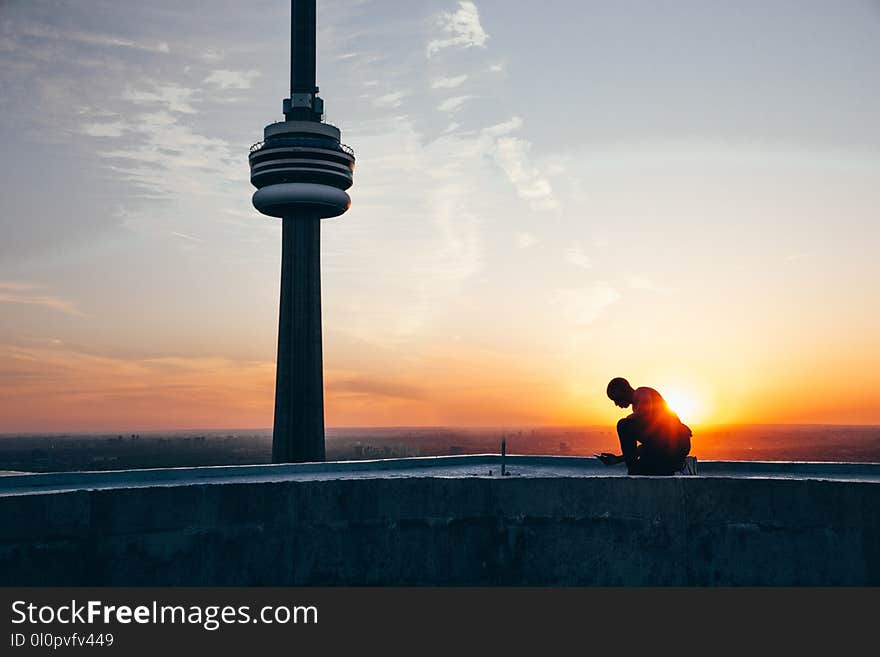 Silhouette of a Man Sitting Near Black Tower Near Body of Water during Sunset