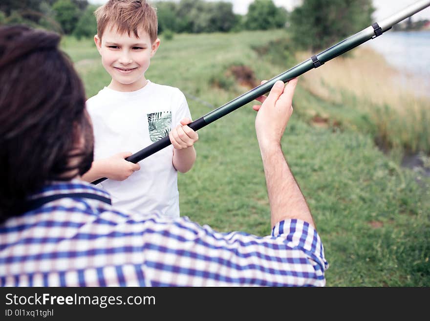 A picture of happy son standing in front of his dad and holding long fish-rod. Guy is holding it a bit. They are