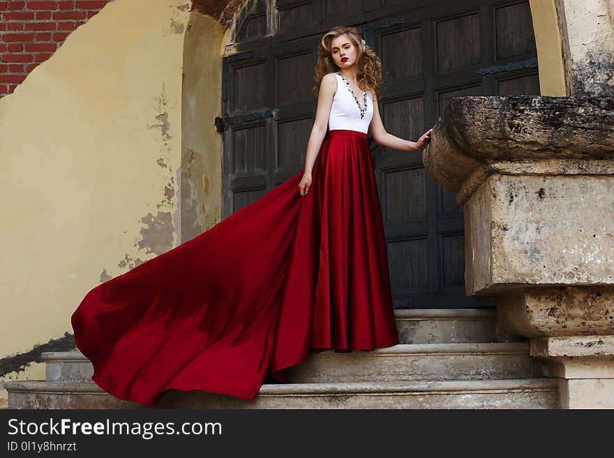 Young beautiful woman standing at stairway and posing in evening dress with white top and red flying dress. Fashion portrait of girl with hairstyling. Young beautiful woman standing at stairway and posing in evening dress with white top and red flying dress. Fashion portrait of girl with hairstyling