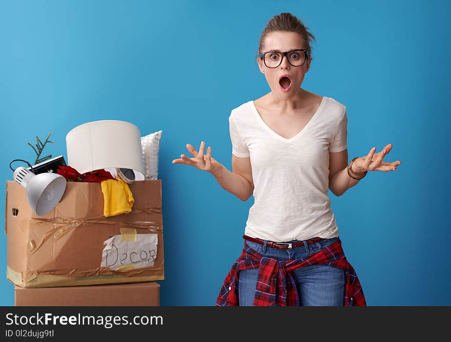 Shocked modern woman in white shirt with an untidy cardboard box in the background isolated on blue. Shocked modern woman in white shirt with an untidy cardboard box in the background isolated on blue