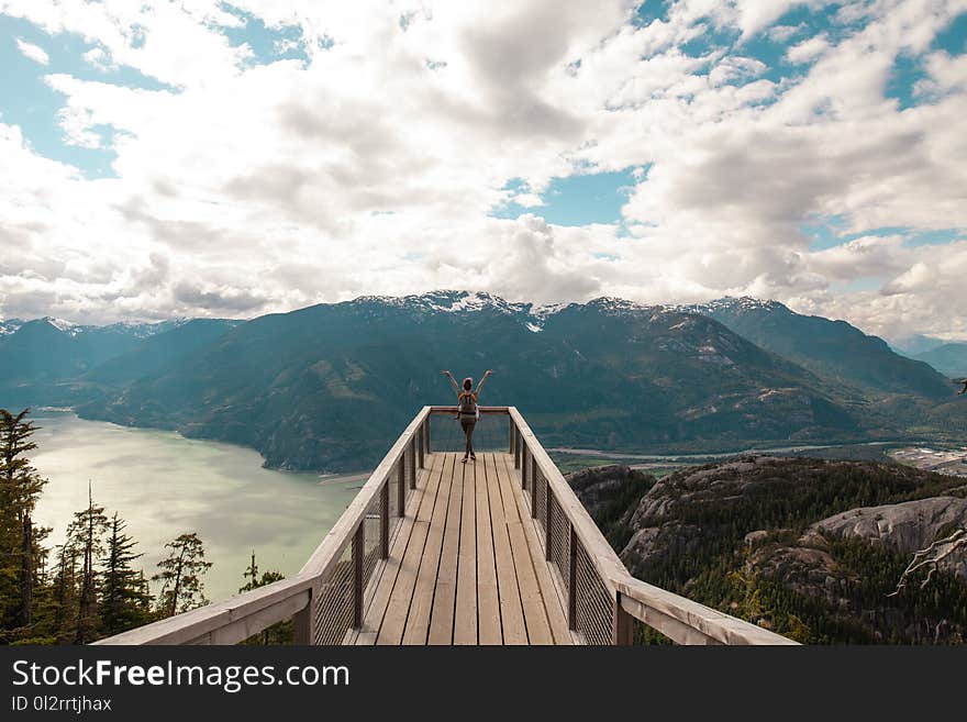 Person Standing With Hands on Air on Brown Wooden Dock With Overlooking View of Lake Under White Clouds and Blue Sky