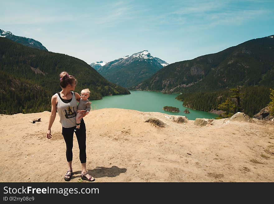 Woman Wearing Grey Tank Top Carrying Baby in Distant of Lake Between Mountains