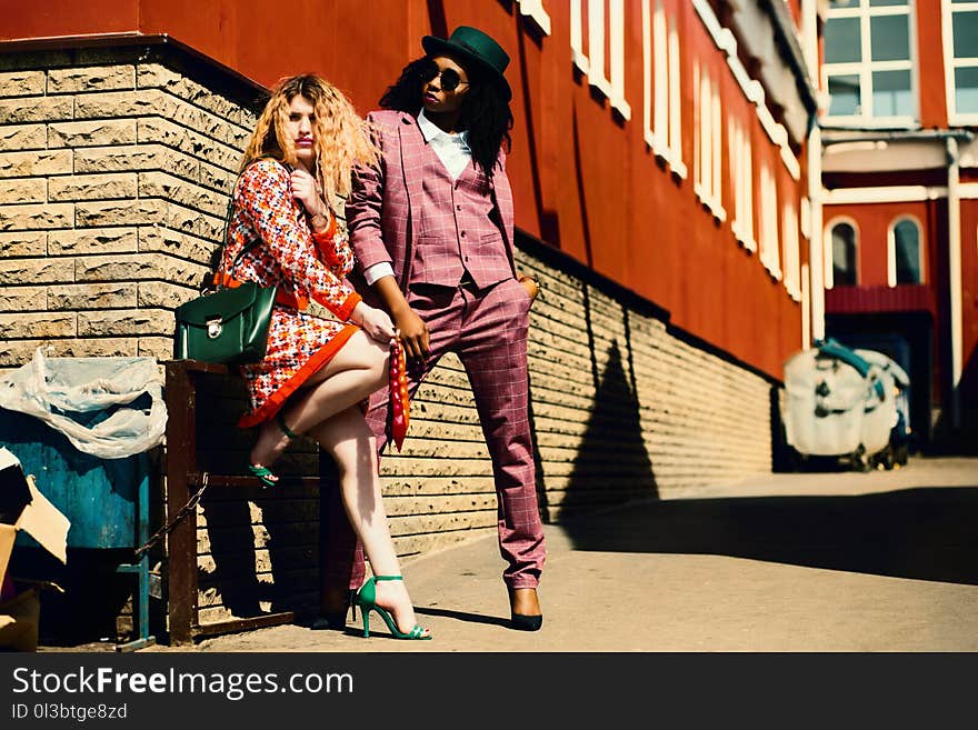 Shallow Focus Photography of Woman in Orange Dress Leaning on Brown Wall Beside Man in Purple Suit Jacket