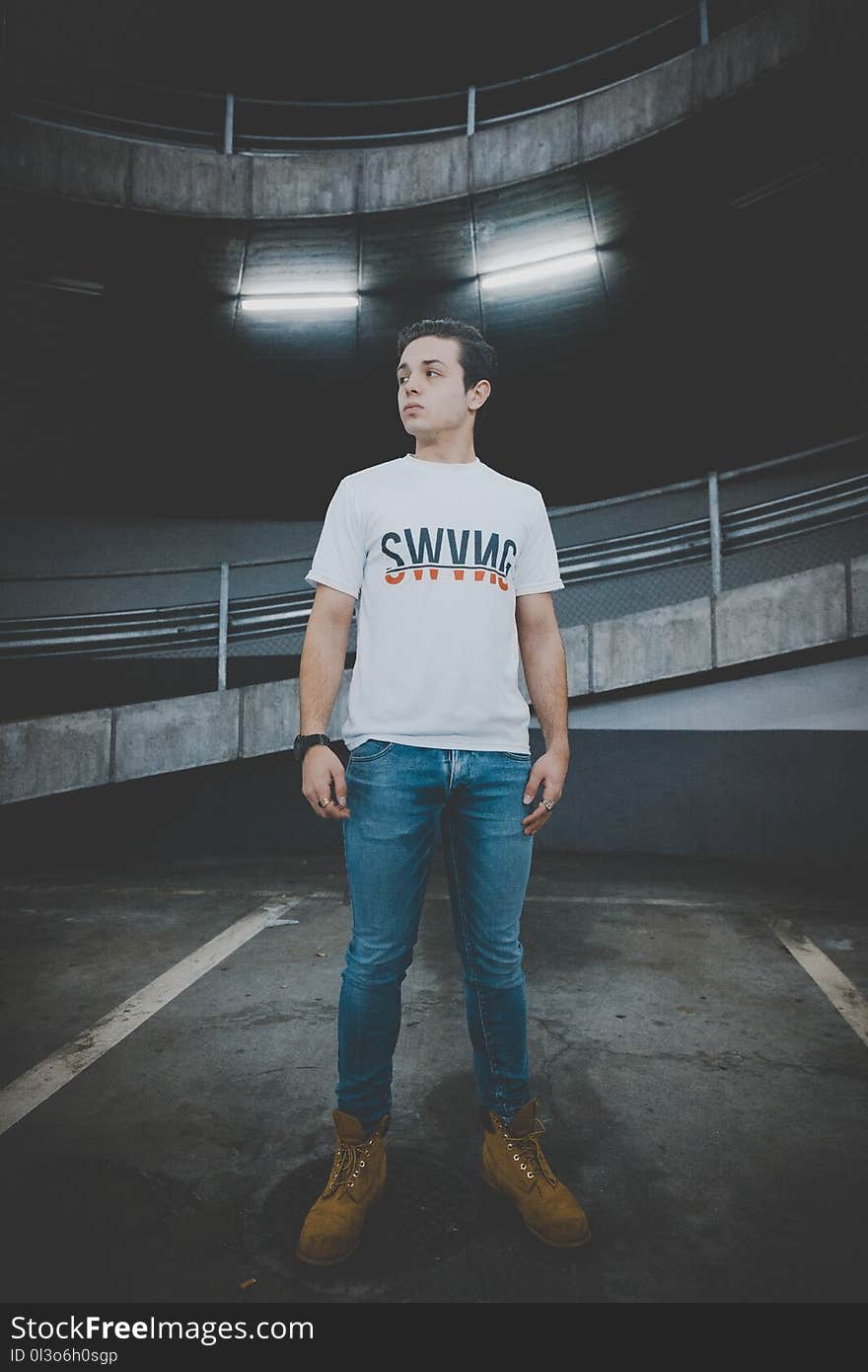 Man Wearing White T-shirt and Blue Jeans Standing on Black Concrete Floor